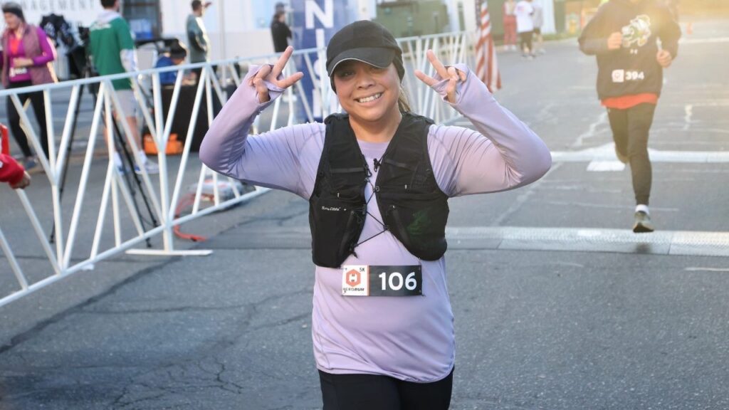 Young woman in purple shirt holds up peace signs to the camera as she crosses the race finish line.