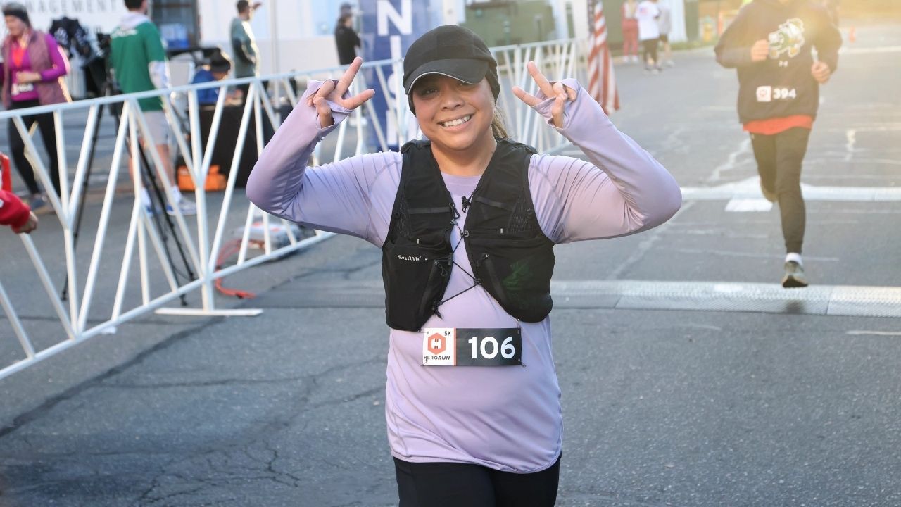 Young woman in purple shirt holds up peace signs to the camera as she crosses the race finish line.